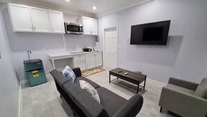 A modern living room with a sofa, a coffee table, and a TV. There is a kitchen area with cabinets and a microwave.