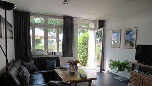 Un salón moderno con un sofá de cuero negro, una mesa de centro de madera y un televisor sobre un mueble de madera. Amplias ventanas dejan entrar luz natural y ofrecen vistas del exterior.
