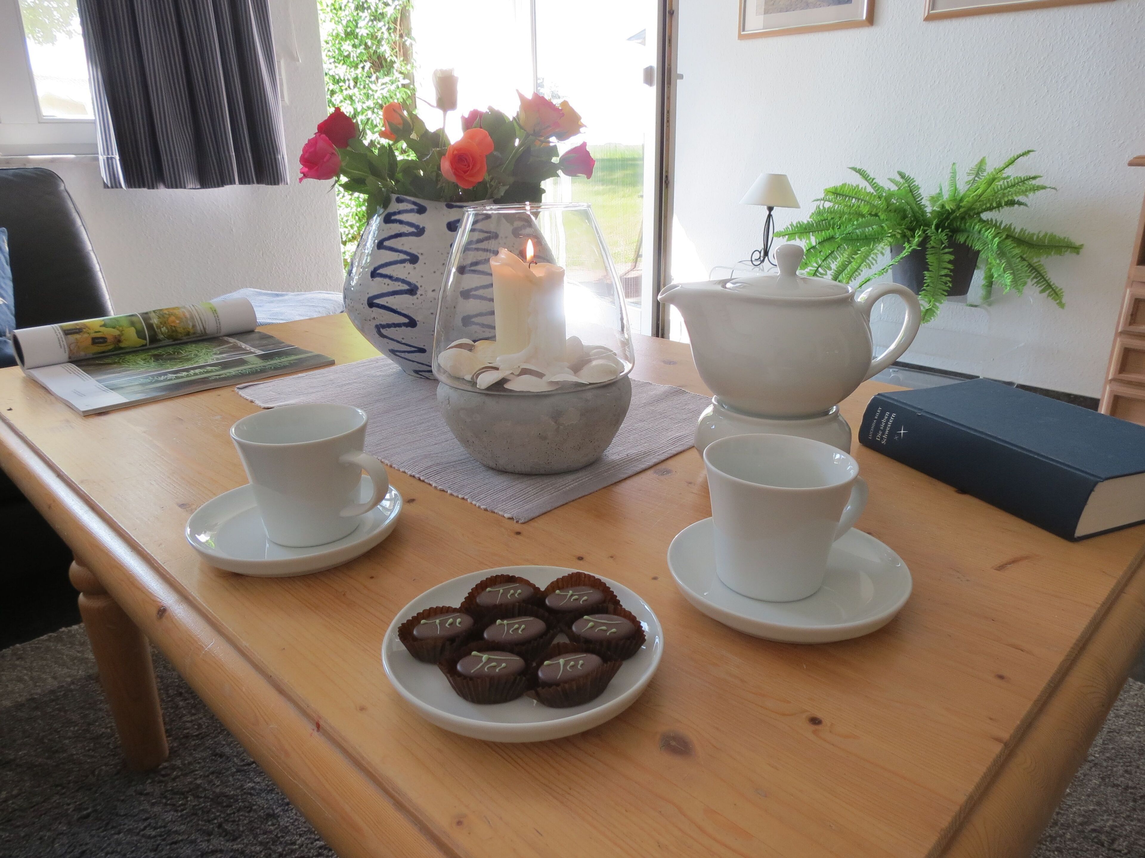 A wooden table with a tea set, a bowl of cookies, a vase with flowers, a teapot, and a book.