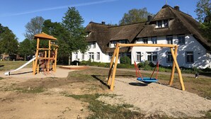 Un parque infantil con columpios y un tobogán, una casa con techo de paja, y un cielo azul despejado.