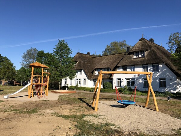 Leikkikenttä, jossa on keinut ja liukumäki, olkikatolla varustettu talo ja kirkkaan sininen taivas.