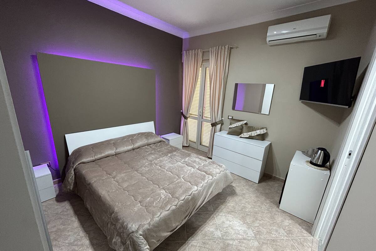 A bedroom with a bed, a bedside table, a dresser, a mirror, a TV, and an air conditioner.