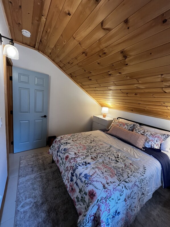 A bedroom with a sloped ceiling, a bed with floral bedding, a nightstand with a lamp, and a door. - Valhalla!! (Ludlow)