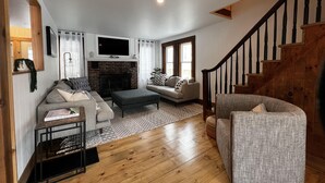 A cozy living room with a fireplace, wooden flooring, a sofa, a chair, and a staircase. - Valhalla!! (Ludlow)