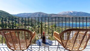 A balcony with wicker chairs and a table with a bottle and glasses, overlooking a mountainous landscape and a body of water. - Villa Naya Desimi in Desimi (Lefkada)