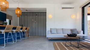 A modern living room with a sofa, a dining area with chairs, and a kitchen in the background. - Villa Naya Desimi in Desimi (Lefkada)