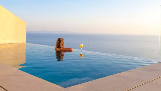 Una persona relajándose en una piscina infinita transparente, sosteniendo un vaso de bebida, con vistas al mar tranquilo.