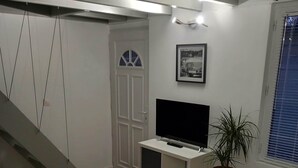 Een knus leefruimte met een trap, een televisie op een witte kast, een klein tafeltje met twee stoelen en een potplant.