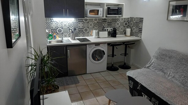 Een compacte keuken met een wasmachine, een magnetron en een broodrooster. Er zijn twee barkrukken en een kleine tafel.
