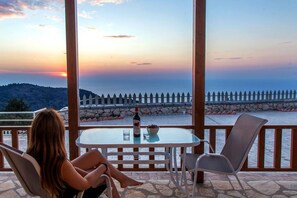 A woman sitting at a table with a bottle of wine and a bowl of grapes on a balcony overlooking a scenic view with a stone wall and a wooden fence.