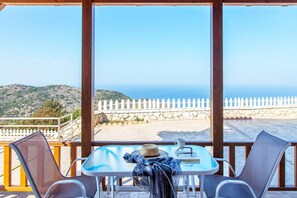 Varanda com mesa e cadeiras, vista para uma praia com cerca branca e parede de pedra.