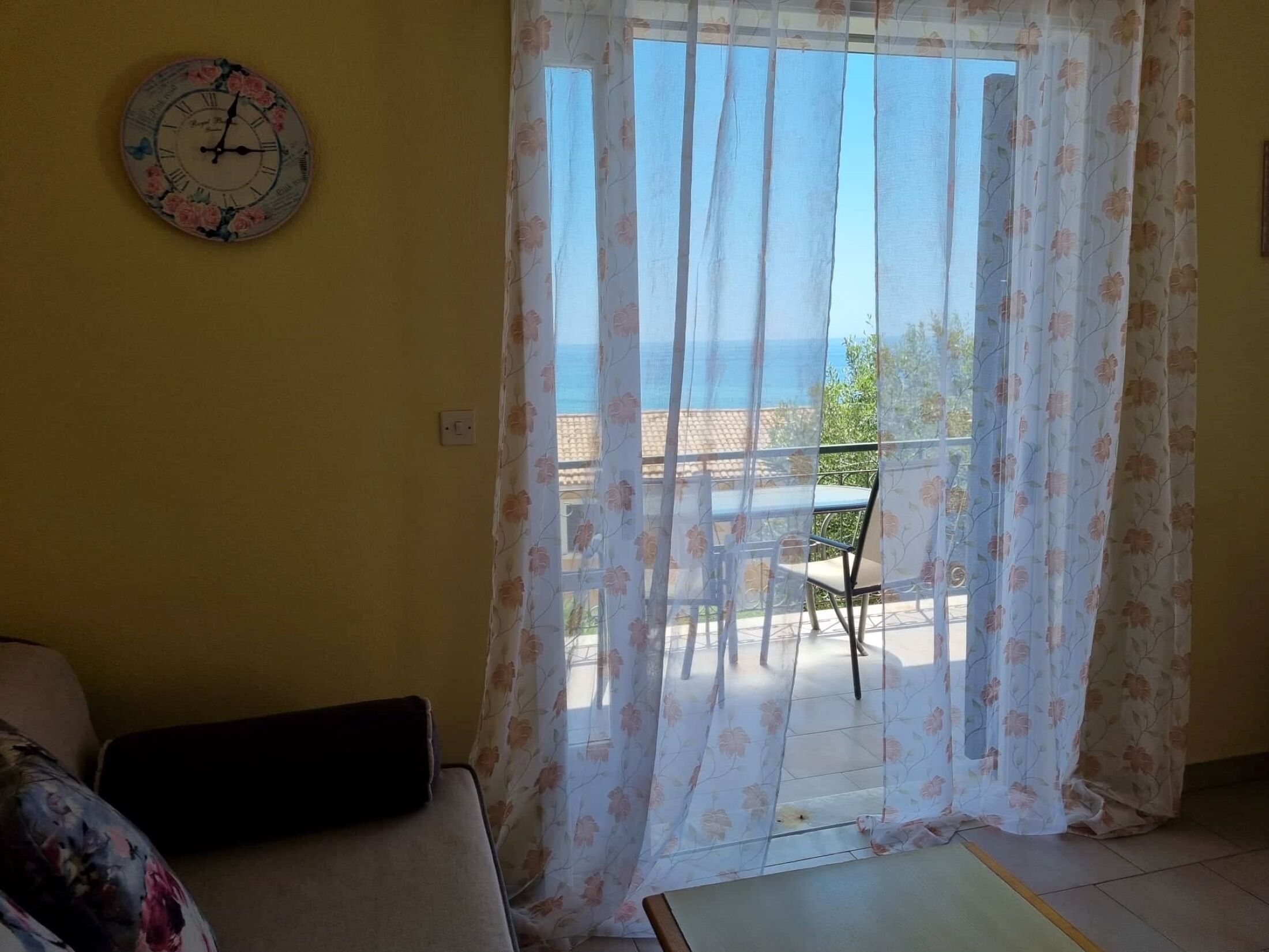 Een kamer met een bed, een stoel, een tafel, een klok en uitzicht op de zee door een raam met bloemen gordijnen.