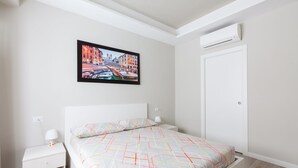 A bedroom with a bed, a nightstand, a wall-mounted air conditioner, and a framed picture of a cityscape. - A Passino from St. Peter's (Roma)