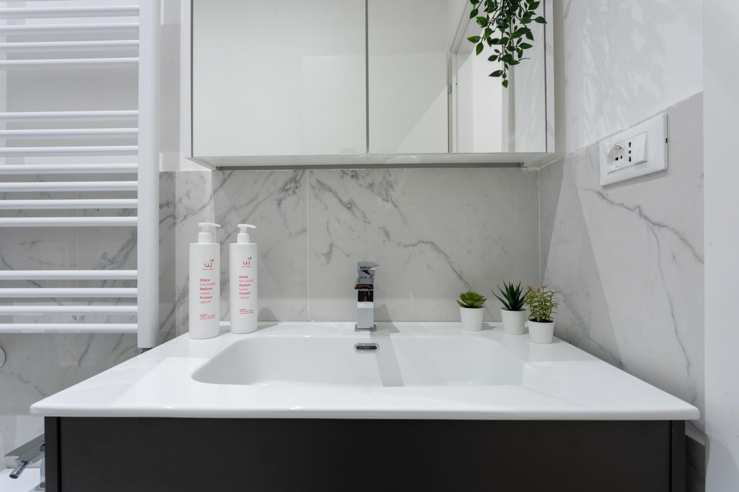 Ein modernes Badezimmer mit Waschbecken, Marmor-Spritzschutz und einem Schrank darüber. Auf der Ablagefläche befinden sich zwei kleine Topfpflanzen.