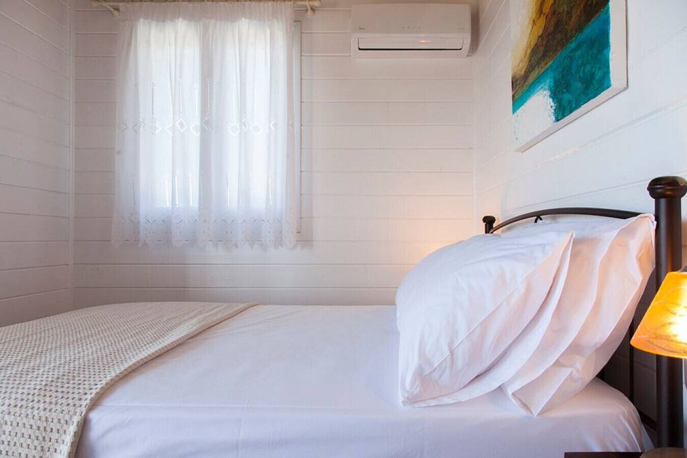 A bedroom with a bed, a window with sheer curtains, an air conditioning unit, and a painting on the wall.