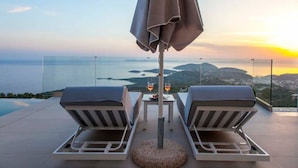 A terrace with two lounge chairs, a table, and an umbrella overlooking a scenic view of the sea and hills. - Syvota 5 Sunset 1B Valentina (Igoumenitsa)