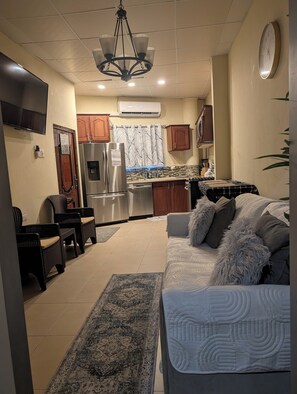 A modern living room with a sofa, a coffee table, and a TV. There is a kitchen area with a refrigerator, stove, and cabinets.