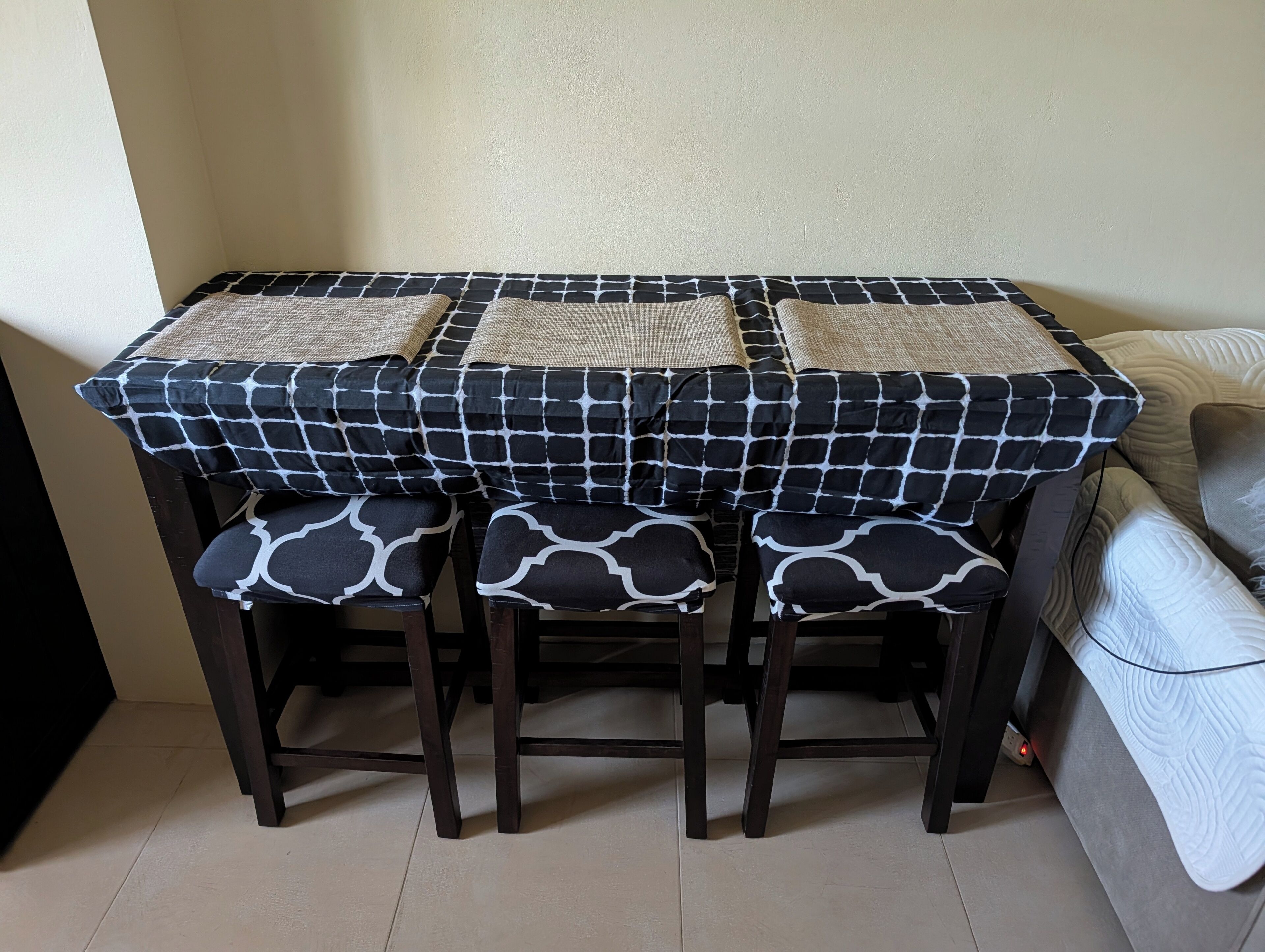 A small dining table with a black and white checkered tablecloth, three matching chairs, and a beige cushion on each chair.