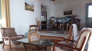 Een woonkamer met een glazen salontafel, twee fauteuils, een rieten mand en een metalen sculptuur.