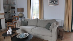 Een woonkamer met een beige bank, een klein rond tafeltje en een salontafel. Er is een boekenkast met decoratieve items en een lamp. Een schilderij hangt aan de muur, en er is een raam met gordijnen te zien.