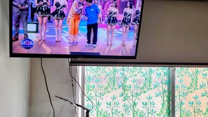 A television mounted on a wall displaying a show with a group of people on a stage. Below the TV, there is a microwave, a remote control, and a mug on a coaster. - Cuarto Privado a 10 Minutos del Parque Kennedy (Lima)