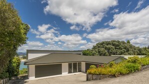 A two-story house with a garage, surrounded by greenery and a clear sky. - Pohutukawa on Paku - Tairua Holiday Home (Tairua)