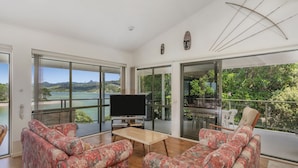 A modern living room with a large TV, floral sofas, and a view of a lake and mountains. - Pohutukawa on Paku - Tairua Holiday Home (Tairua)