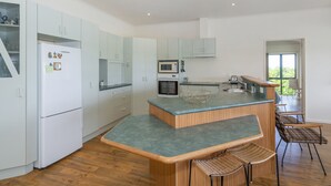 A modern kitchen with white cabinets, a central island, a microwave, oven, and bar seating with chairs. - Pohutukawa on Paku - Tairua Holiday Home (Tairua)