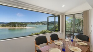 A dining area with a table set for four, overlooking a scenic bay with water and greenery. - Pohutukawa on Paku - Tairua Holiday Home (Tairua)