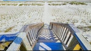 Ein hölzerner Steg führt zu einem Strand mit weißem Sand und spärlicher Vegetation.