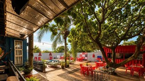 An outdoor patio area with red tables and chairs, a covered seating area, and a view of a red building in the background. - Tetris Container Hostel (Foz do Iguaçu)