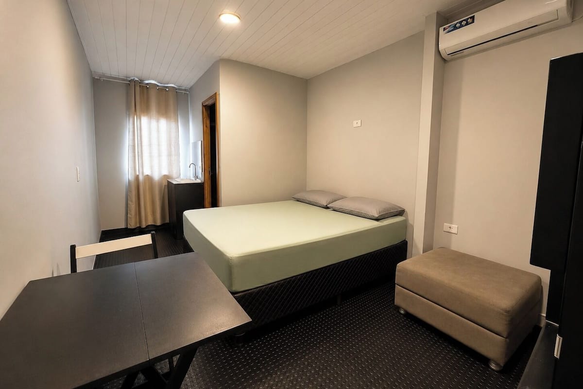 A small, compact room with a single bed, a small table, a bench, an air conditioning unit, and a window with curtains.