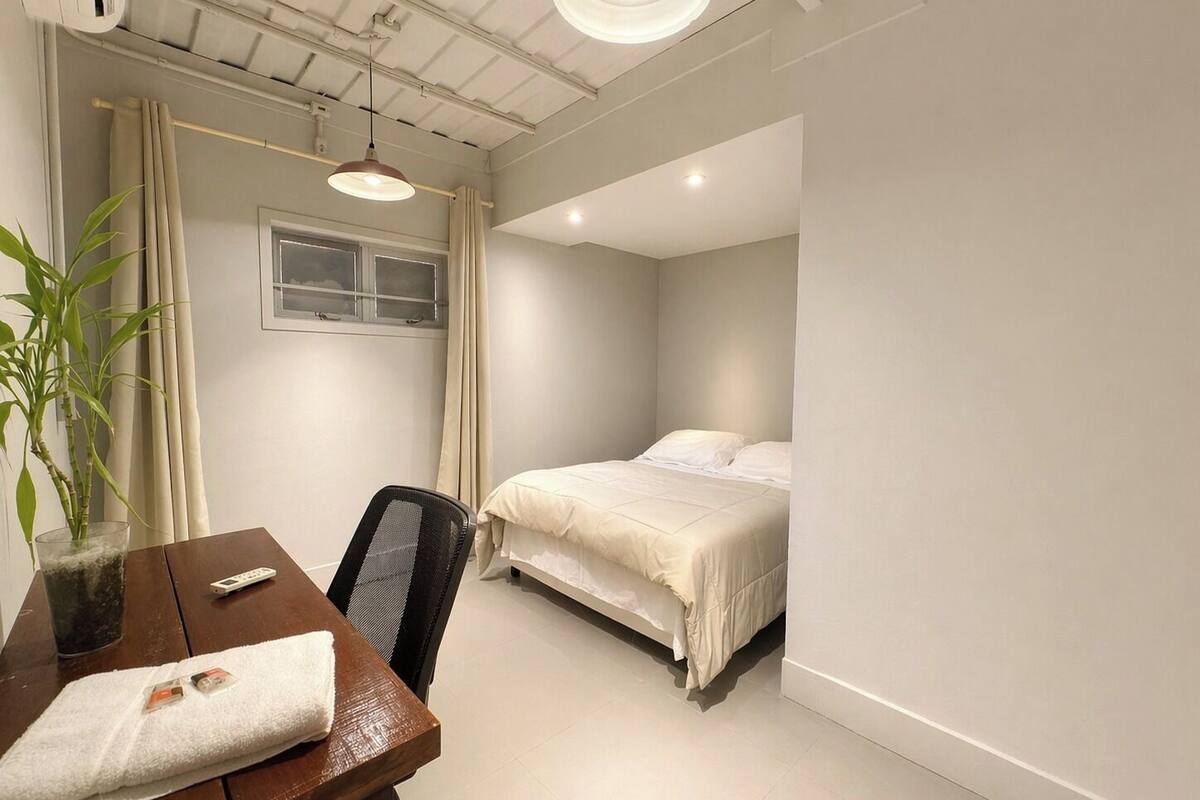 A bedroom with a bed, a desk with a chair, a potted plant, and a window.