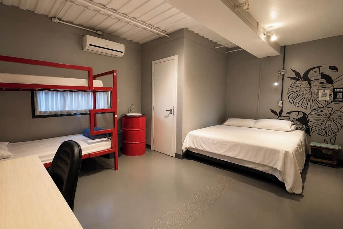 A dormitory room with bunk beds, a desk, a chair, a red barrel, a door, and a wall with a plant mural.