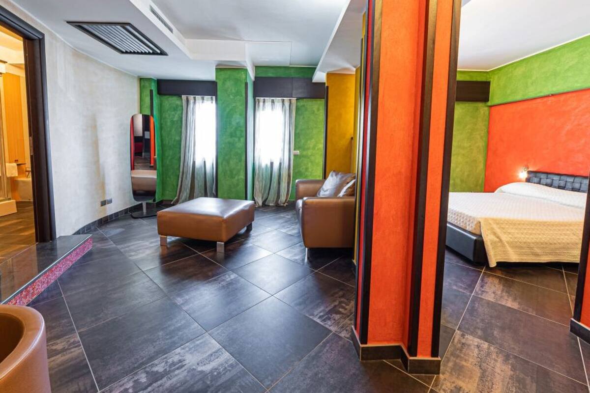A modern hotel room with a large bed, a seating area with a sofa and ottoman, a small table, and a view of the hallway.