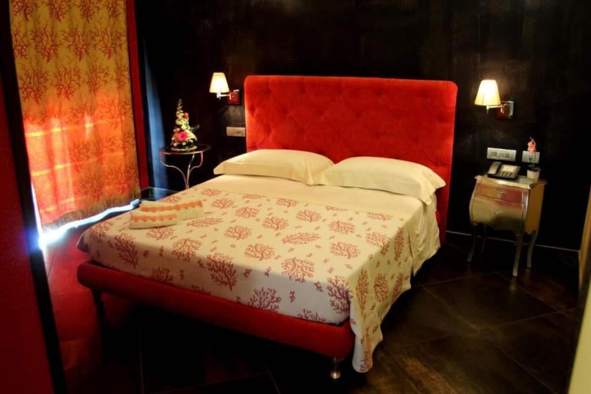 A hotel room with a red upholstered bed, a small bedside table with a lamp, and a decorative item on the nightstand.
