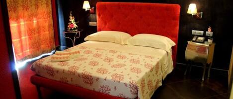 Habitación de hotel con una cama tapizada en rojo, una mesita de noche pequeña con una lámpara y un objeto decorativo en la mesita.