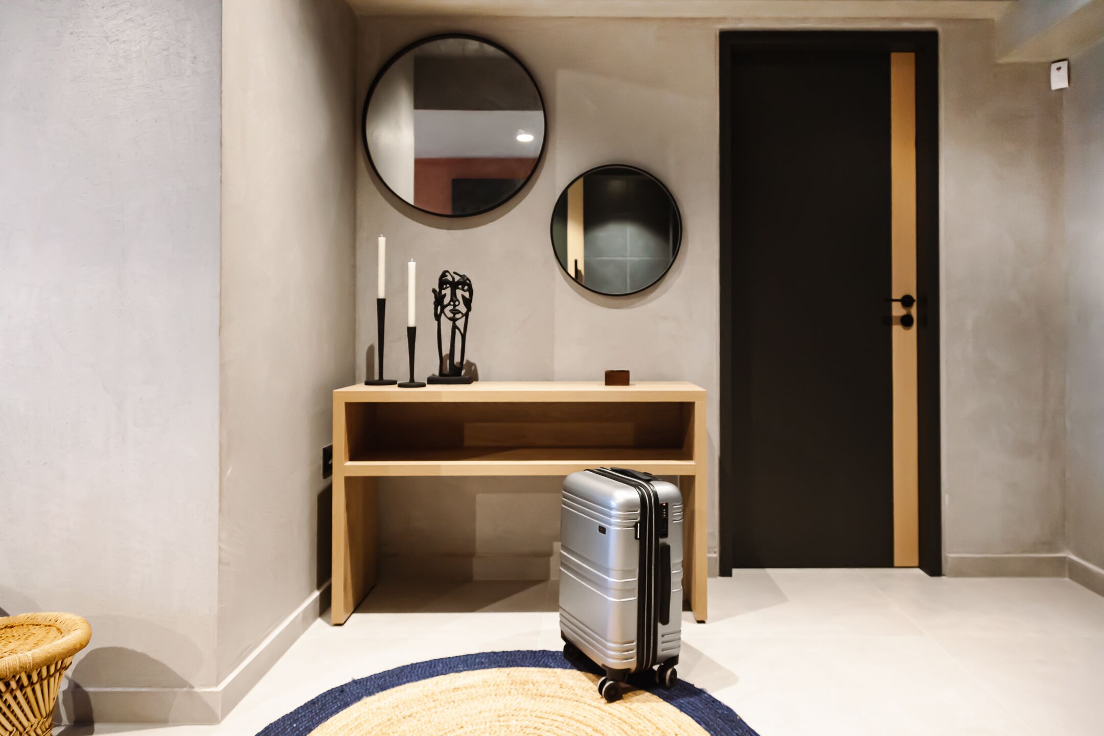 Een hal met een houten console tafel, een ronde spiegel, een deur en een koffer op een geweven vloerkleed.