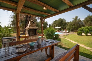 An outdoor patio with a stone table set for dining, a stone fireplace, and a pool in the background.