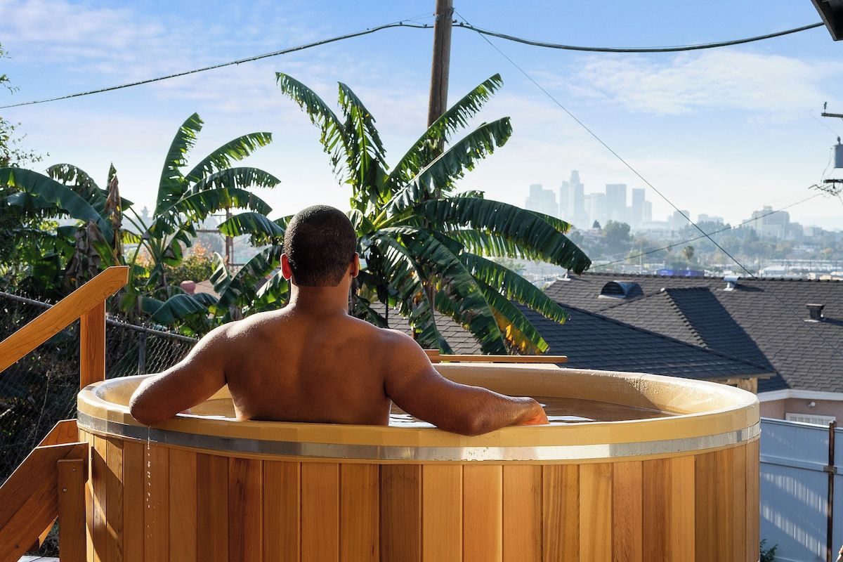 Iemand ontspant in een houten jacuzzi, met een stadsachtergrond op de achtergrond.