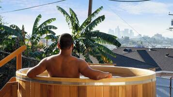 Iemand ontspant in een houten jacuzzi, met een stadsachtergrond op de achtergrond.