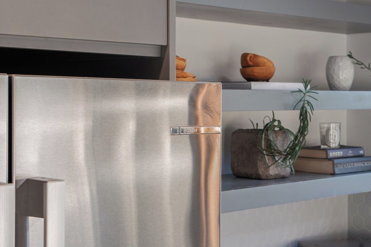 A stainless steel Bosch refrigerator in a modern kitchen with a shelf above containing decorative items and books.
