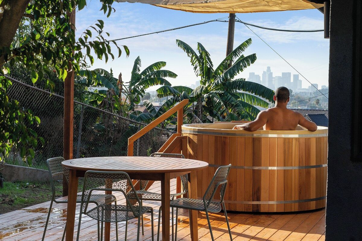 Un bain à remous en bois, avec la silhouette de la ville en arrière-plan, entouré de verdure et doté d'une terrasse en bois avec des chaises.