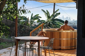 A wooden hot tub with a city skyline in the background, surrounded by greenery and a wooden deck with chairs.
