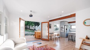 Un salon moderne avec un canapé, une table basse et un tapis à motifs. On y trouve également une table à manger en bois avec des chaises, ainsi qu’un coin cuisine équipé d’un réfrigérateur et de tabourets de bar.