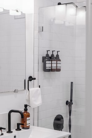 A modern bathroom with a white tiled wall, a glass shower enclosure, a black faucet, a white sink, and toiletries.