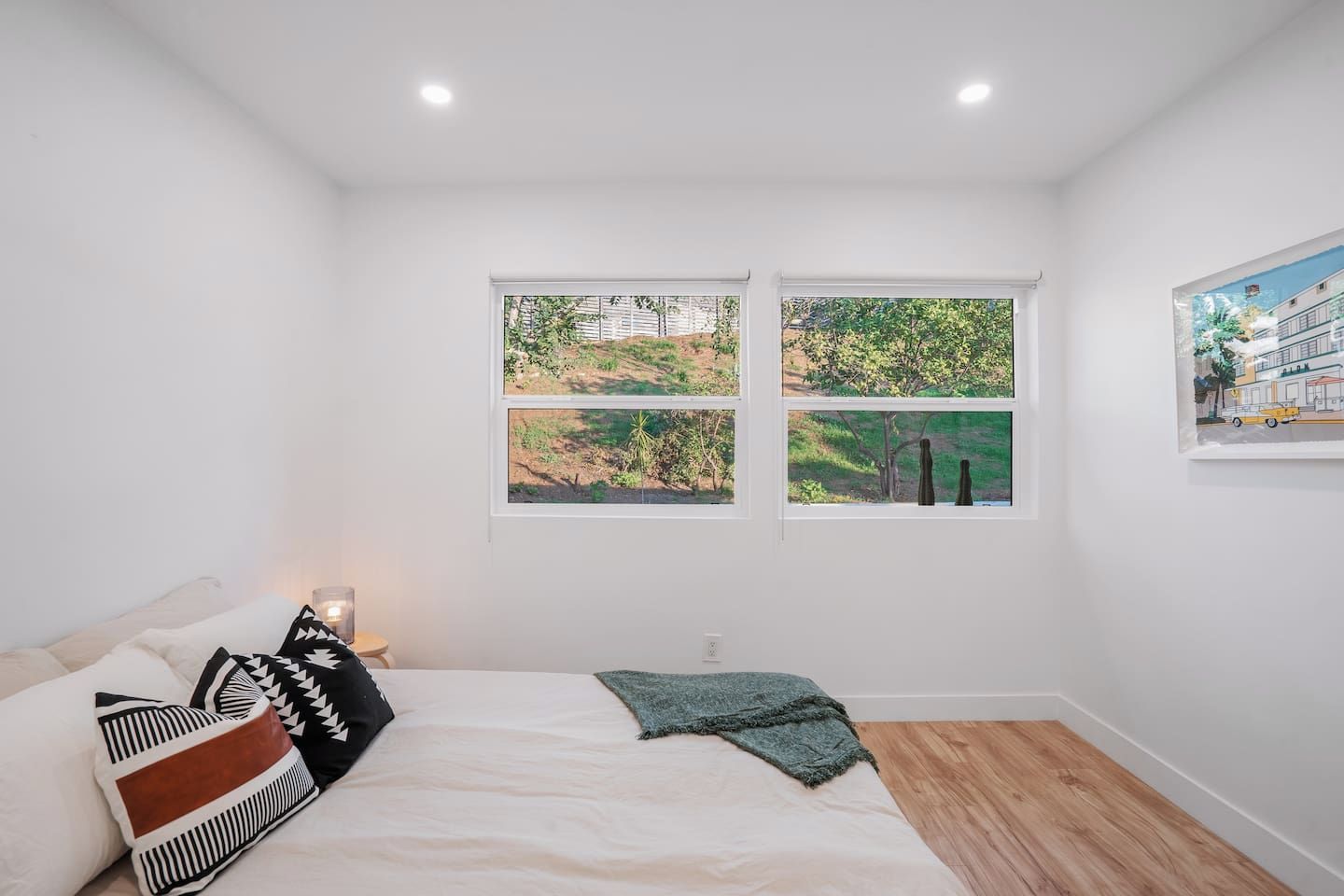 A bedroom with a bed, pillows, a bedside table with a lamp, two windows with views of greenery, and a framed picture on the wall.