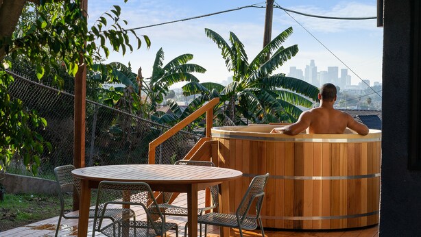 Un bain à remous en bois, avec la silhouette de la ville en arrière-plan, entouré d’une végétation luxuriante et agrémenté d’une terrasse en bois avec des chaises en métal.