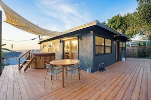 A modern outdoor deck with a wooden table and chairs, a hot tub, and a small building with large windows.