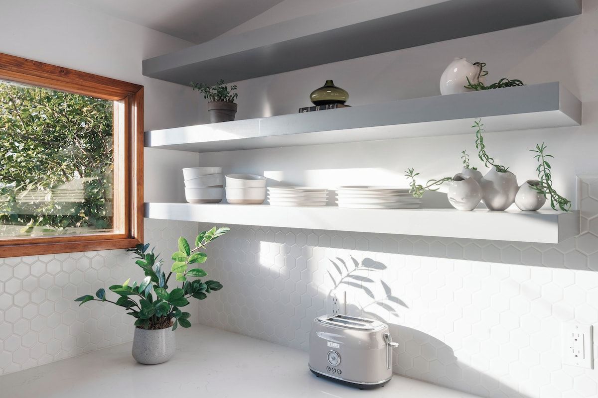 A kitchen with white hexagonal tiles, white countertops, a toaster, and shelves with kitchenware and plants.